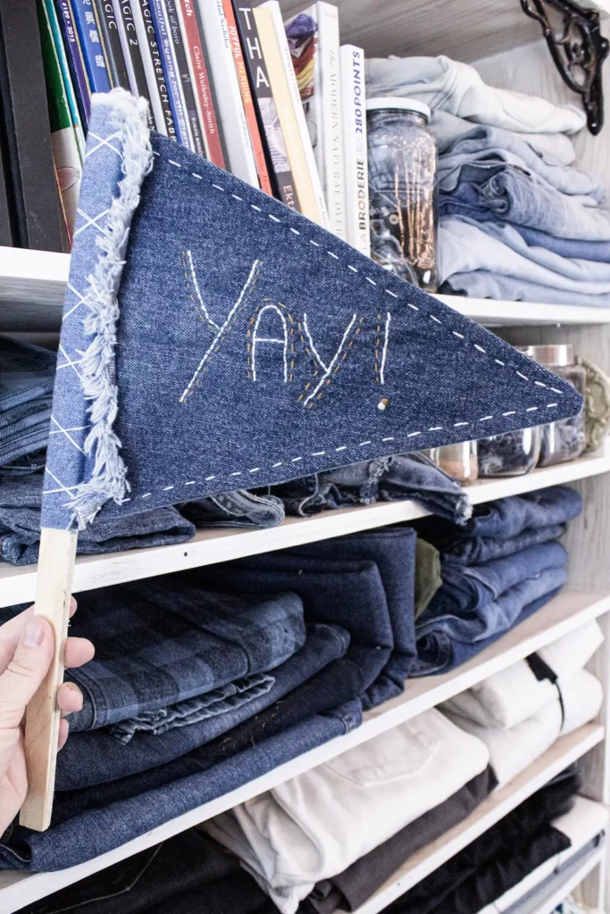 Drapeau confectionné à partir de denim récupéré et brodé du mot « Yay! », devant une étagère de vieux jeans dans l’atelier Textilab.