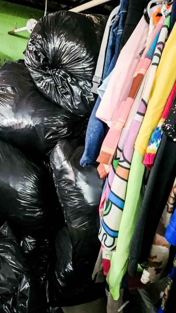 Vêtements colorés dans une friperie, avec des sacs de dons empilés jusqu’au plafond, illustrant l’accumulation de textiles