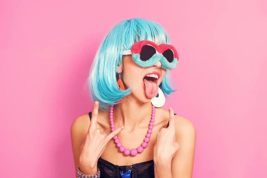Jeune femme portant une perruque bleue et des lunettes roses et bleues, faisant une grimace dans une ambiance ludique et colorée